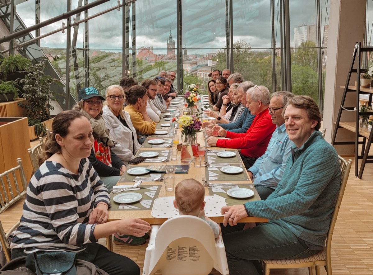 A group of about twenty travellers of several generations seated along one long dinner table, with a city skyline visible through the glass wall behind them