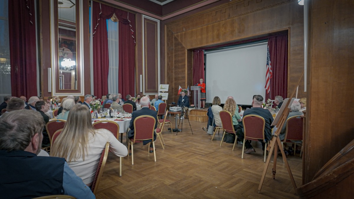 A commemorative dinner in a wood-panelled hall, with American and Polish flags beside a speaker at the lectern