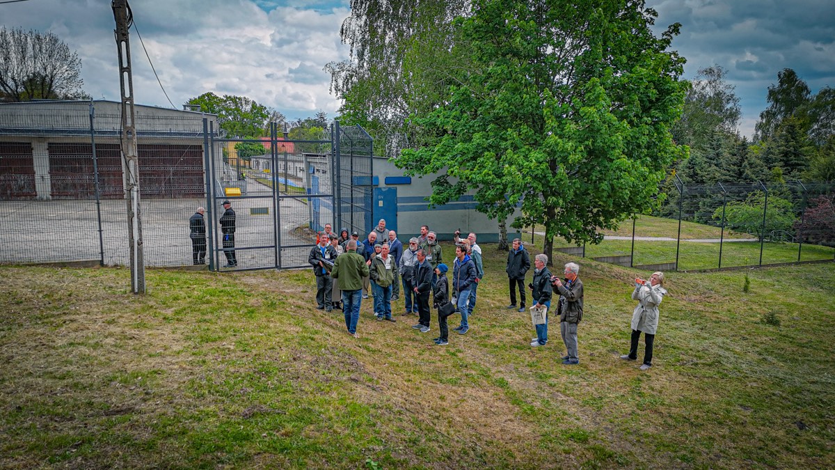 Aerial view of the group standing on the grass outside the fenced grounds of the former Oflag 64 camp site in Szubin